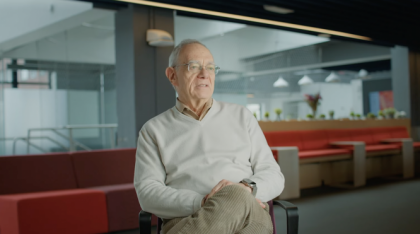 Rafael Reif wears a light-colored sweater and sits on a chair. The background has red couches, modern office furniture, and large windows.