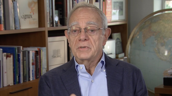 Rafael Reif speaks in front of a bookshelf filled with various books. A large globe is visible to the right, partially obscured. The background includes framed pictures and additional bookshelves.